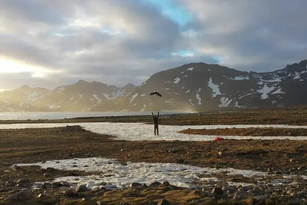 یک پژوهشگر دست خود را بهسوی هوا دراز میکند تا یک پهپاد کوچک پرتاب کند. پسزمینه یک منظرهٔ کوهستانی از جزیره است