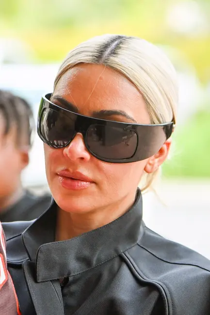 کیم کارداشیان با عینک‌های بزرگ و لباس شیک با یقه بلند در فضای باز