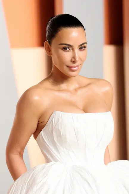 کیم کارداشیان در لباس سفید با بافت و بدون آستین، در مقابل پس‌زمینهٔ هندسی ایستاده