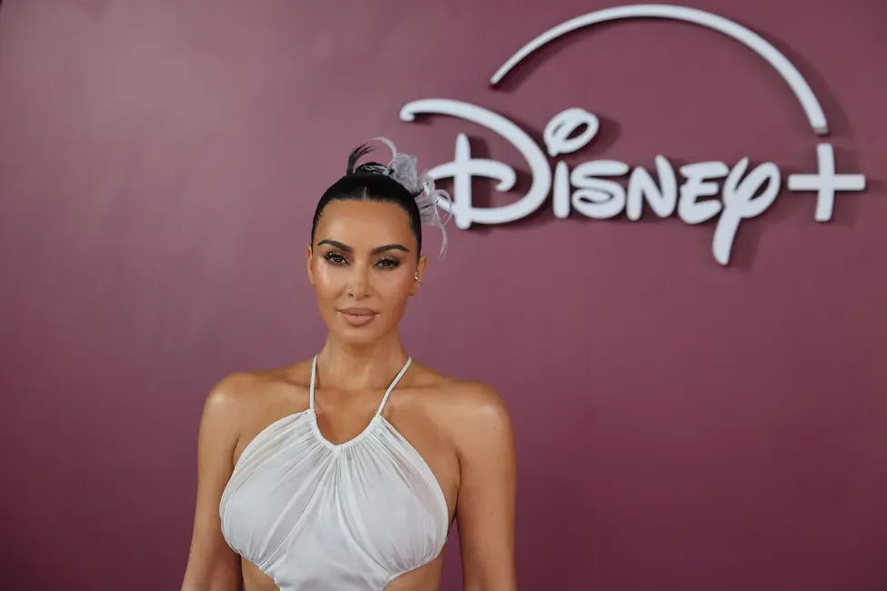 کیم کارداشیان در لباس سفید شیک بدون آستین، در مقابل لوگوی Disney+ روی فرش قرمز ایستاده
