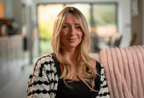 الن، زنی با موهای بلند بلوند، بر روی یک مبل نشسته و به دوربین لبخند میزند