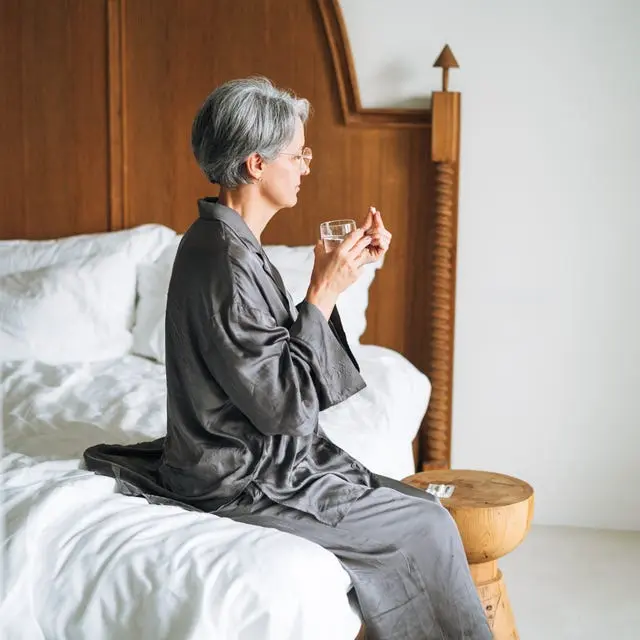 زن میانسال زیبا با موهای خاکستری که در خانه بر روی تخت نشسته، آب مینوشد و قرص میخورد