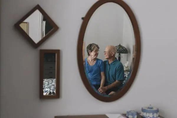 ری هستر روی تخت کنار همسرش ساندی نشستهاند و هر دو به هم لبخند میزنند؛ این صحنه در آینهای بر روی دیوار اتاق خواب منعکس شده است.