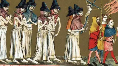 گتی: هشت نفر پوشیده در رختهای سفید و کلاههای نوکدار، با پاهای عریان در کاروانگردی پشت سر گروهی از سه مرد با لباسهای رنگارنگ میپیمایند. این مردان پرچمها را حمل میکنند.