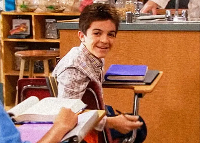 پسر جوانی که در کلاس نشسته، لبخند میزند و قلم در دست دارد؛ مرتبط با بهروزرسانی سلامت پدر ستارهٔ نیکلودئون تیلور چیس.