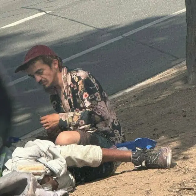 مردی که گمان میرود پدر ستارهٔ نیکلودئون تیلور چیس باشد، روی پیادهرو نشسته و نشانههای بیخانمانی و مشکلات سلامتی را نشان میدهد.