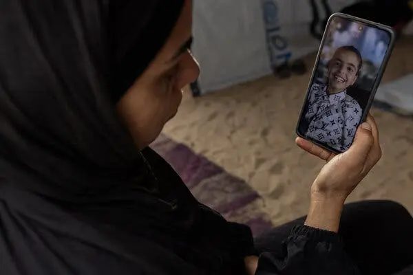 زنی با پوشش سر سیاه به سمت گوشی هوشمند نگاه می‌کند. صفحه‌نمایش کودک با دهان باز و لبخند پهن، در پیراهن سفید طرح‌دار، را نشان می‌دهد.